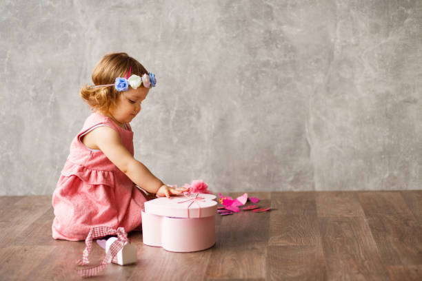 Adorable baby girl opening a gift Portrait of adorable baby girl with flower crown sitting on the floor against gray wall, opening a gift box. floral crown photos stock pictures, royalty-free photos & images
