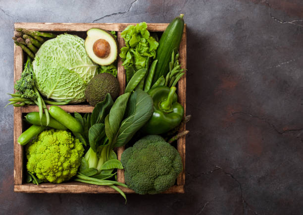 istock Assorted green toned raw organic vegetables in wooden box on dark background. Avocado, cabbage, cauliflower and cucumber with trimmed and mung beans and pepper and broccoli with sparagus tips. 1142446573