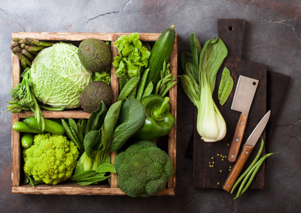 istock Assorted green toned raw organic vegetables in wooden box on dark background. Avocado, cabbage, cauliflower and cucumber with trimmed and mung beans and chopping board with knife. 1142458748