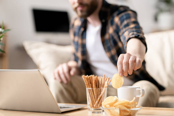 Having snack during work Young man taking potato chip out of glass bowl while sitting on sofa in front of laptop on table and having snack snack stock pictures, royalty-free photos & images