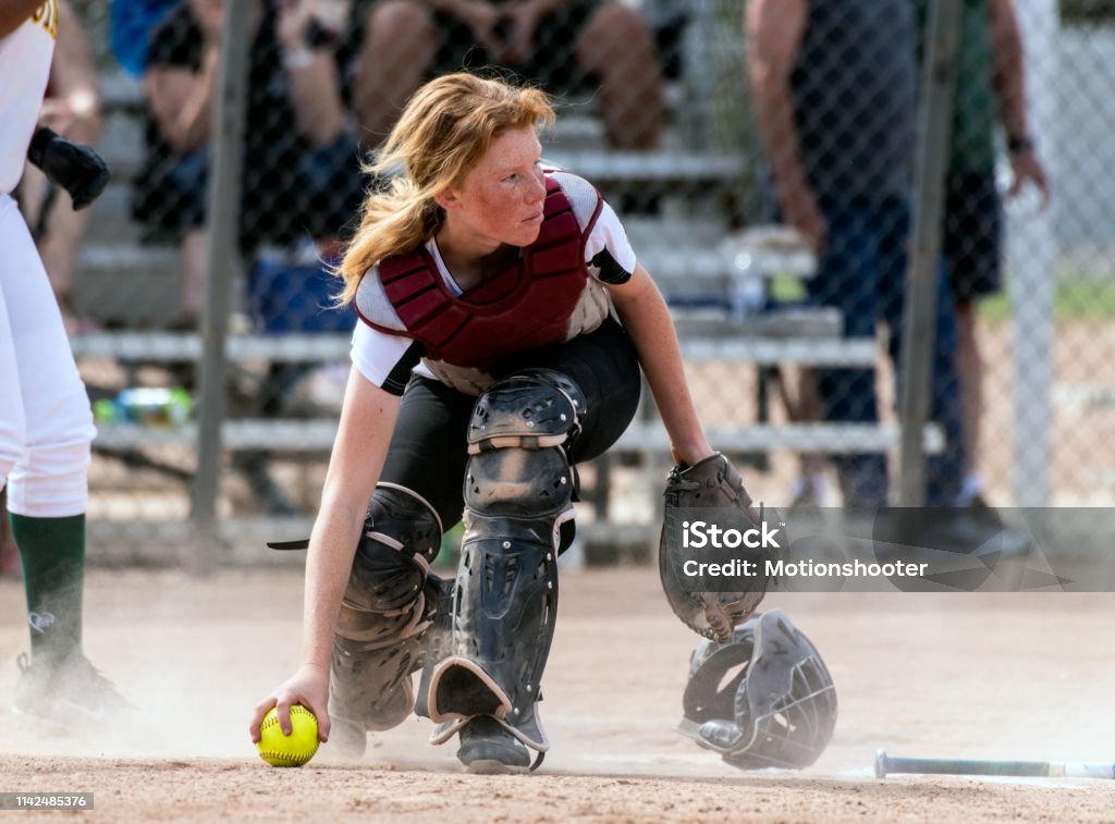Hustling in a cloud of dust Skilled softbaall catcher with red hair and protective gear gaining a grip on the loose ball while looking up field. High School Stock Photo