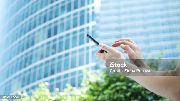 女性の手はガラスのビジネスセンターと緑の木々の背景にスマートフォンを使用しています - インターネットのストックフォトや画像を多数ご用意 - インターネット, ガラス, クローズアップ