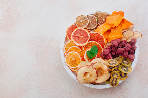 Mixed dried fruit chips in round ceramic plate on beige concrete background with blank space for text. Top view, flat lay.