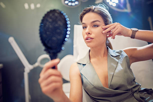 la giovane donna guarda il suo viso allo specchio dopo la procedura di trattamento della bellezza - nose job foto e immagini stock