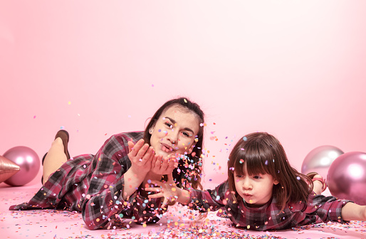 Funny mom and child lie on a pink background. Little girl and mother having fun with confetti. The concept of celebration and fun.