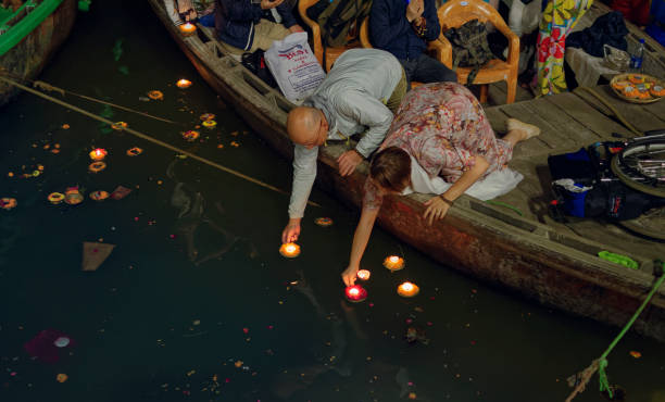 Scene of Dashashwamedh ghat in Varanasi, during evening aarti (puja / religious ritual). Tourists, here two foreigners float diyas (earthen candle, decorated with flower) in the holy Ganges river water, from boats floating on water. This ritual is a form of worshipping the holy Ganges, a sacred river in Hindu religion. Few other tourist visible in background.\n\nVaranasi@ Uttar Pradesh is one the oldest living cities of world and called spiritual capital of India. The city is on the banks of river Ganga. Though the city and its people have adopted new times, but still much of city has preserved the old charm. \nPhoto taken at Varanasi, Uttar Pradesh on 11/17/2018