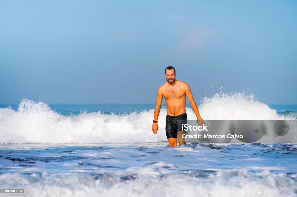 young attractive and happy man with beard and swimming trunks at tropical paradise desert beach alone playful and cheerful in sea water enjoying Summer holidays destination isolated blue 30-39 Years Stock Photo