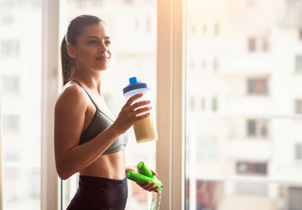 Bodybuilder girl relax after exhausting training, Young athlete drinking sports drink after workout, beautiful woman resting after exercising training. Bodybuilder girl relax after exhausting training, Young athlete drinking sports drink after workout, beautiful woman resting after exercising training, young sports model holding protein shaker and skipping rope around the window. protein stock pictures, royalty-free photos & images