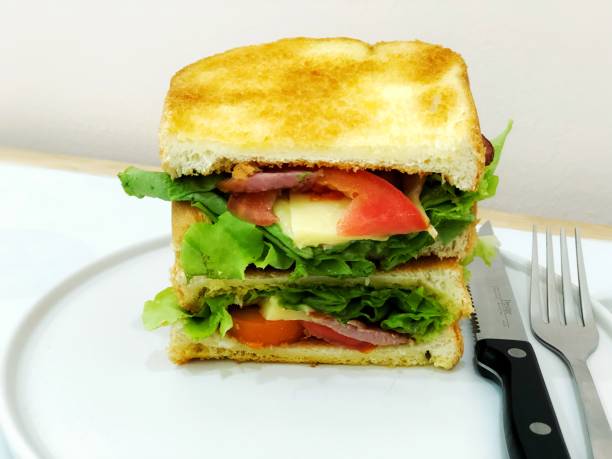 Sandwich toasted bread cheese hamburger, cooked with bacon, cheese, tomato, lettuce and vegetable put on white plate with fork and knife for breakfast or lunch time, selective focus blur background