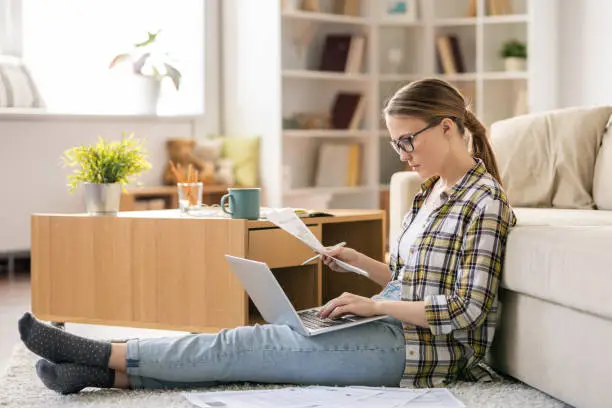 Serious concentrated young woman in glasses sitting on floor in living room and using laptop while checking bills at home