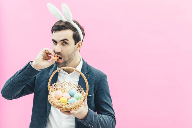 istock Young handsome man standing on a pink background. At the same time holding a basket with Easter eggs with one hand. Another puts carrots in the mouth looking at the camera. 1142633314