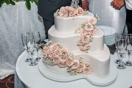 Three-tiered white wedding cake decorated with roses and glasses for champagne on restaurant table ready for bride and groom.