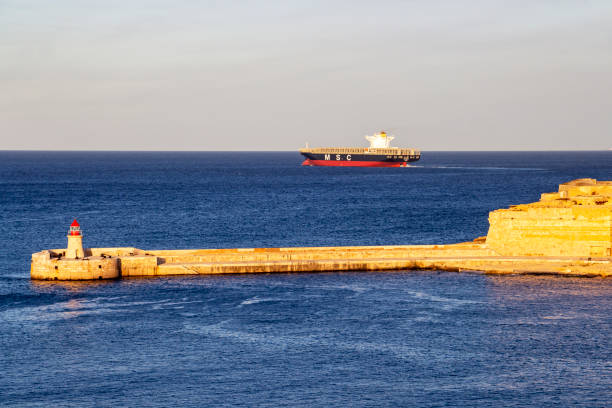 Kalkara, Malta - March 06, 2018: Deep blue seascape with Ricasoli Breakwater Lighthouse at Grand Harbor, Malta and a MSC container ship