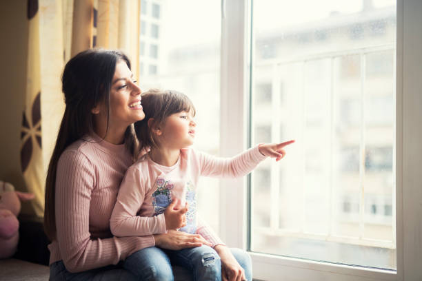mother and daughter together looking out the window at home and smiling happy. - mother holding child pointing imagens e fotografias de stock