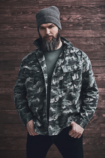 Style. Bearded man in camouflage jacket is looking at camera while standing with his hands in pockets on a wooden background