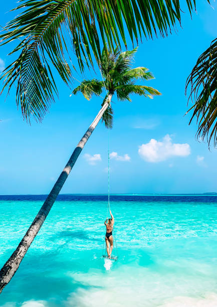 雲の上に 9.モルディブのターコイズブルーの水の上にある鮮やかな緑のヤシの木で、ブランコがあり、スポーティな女の子が黒い水着を身につけています。 - tourism travel travel destinations vertical ストックフォトと画像