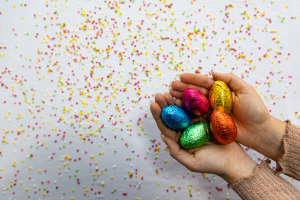 istock Woman hands holding colorful chocolate easter eggs with white background and colorful blurred confetti 1142697653