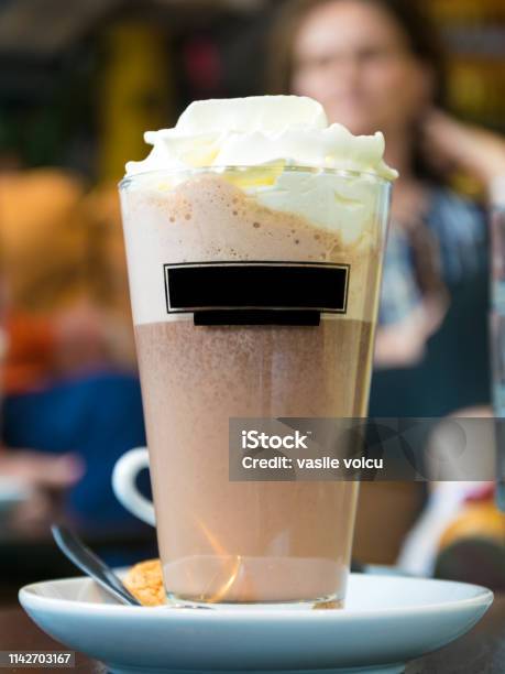 A Cup Of Frappe With Chocolate And Whipped Cream Served In A Transparent Glass Placed On A Saucer With A Lighter With A Person In The Background Stock Photo - Download Image Now