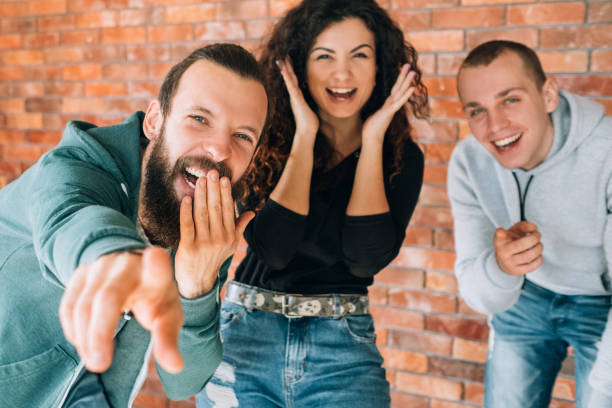 Amused, cheerful millennial mocking. Young emotional people laughing, having fun together. Hipster guy pointing finger at camera.