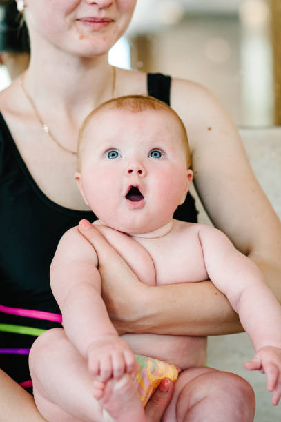 maman tenant bébé fille 6 mois, un bébé dans ses bras à la piscine. la première leçon de natation. préparez-vous à plonger. surpris. avec une bouche ouverte. cheveux roux. close-up. enfant. gardez vos pieds dans les mains - family mouth open vertical mother photos et images de collection