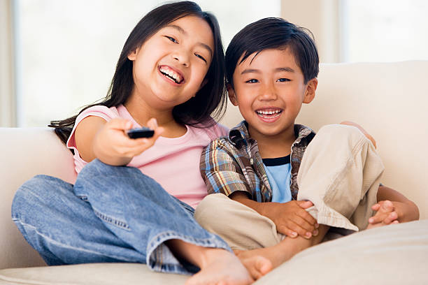 Two young children in living room with remote control smiling Two young children sitting on sofa in living room with remote control smiling asian kids watching tv stock pictures, royalty-free photos & images
