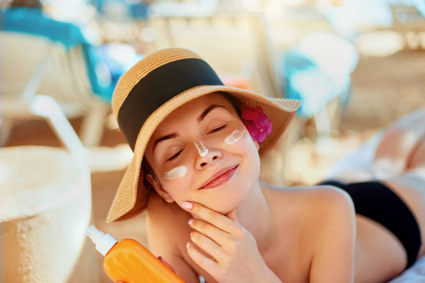 Young woman with sun cream on face holding sunscren bottle on the beach. Female in hat applying  moisturizing lotion on skin. stock photo