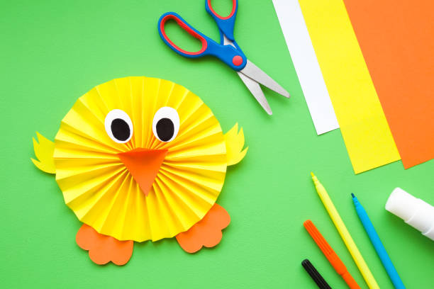 Cute, yellow chick who folded and created from paper for kids. Colorful application paper, scissors, glue stick and color pens on green desk. Top view.
