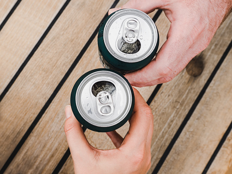 Two can of beer, female and male hands on the background of a beautiful, wooden surface. Top view, close-up. Concept of leisure and travel