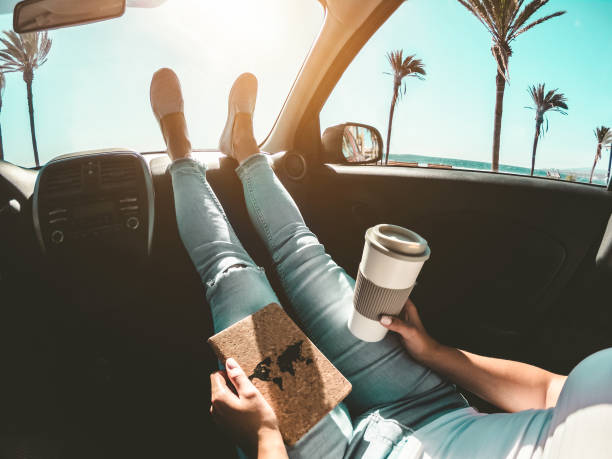 kobieta pijąca filiżankę papieru kawowego wewnątrz samochodu ze stopami na desce rozdzielczej - dziewczyna relaksująca się w auto-podróży czytając książkę podróżną z oceaniczną plażą i palmami w tle - koncepcja podróżnika - skup się na - coffee time zdjęcia i obrazy z banku zdjęć