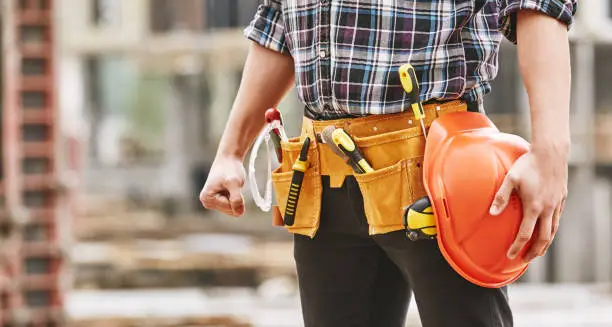 Safe work. Cropped photo of male professional builder with construction tools holding a safety red helmet while standing outdoor of construction site. Industry concept. Building concept. Technology concept.