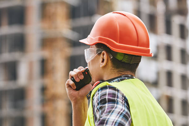 dando instruções. foto cortada do construtor no uniforme de funcionamento e no capacete protetor vermelho que falam ao operador do guindaste pelo walkie talkie ao estar no canteiro de obras. - commercial dock audio - fotografias e filmes do acervo