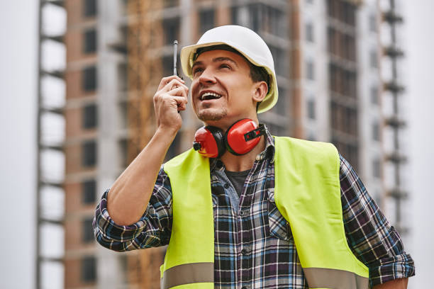 istock Turn left! Cheerful young builder in working uniform, yellow protective helmet talking to crane driver by walkie-talkie, looking up and smiling while standing at construction site. 1142818855
