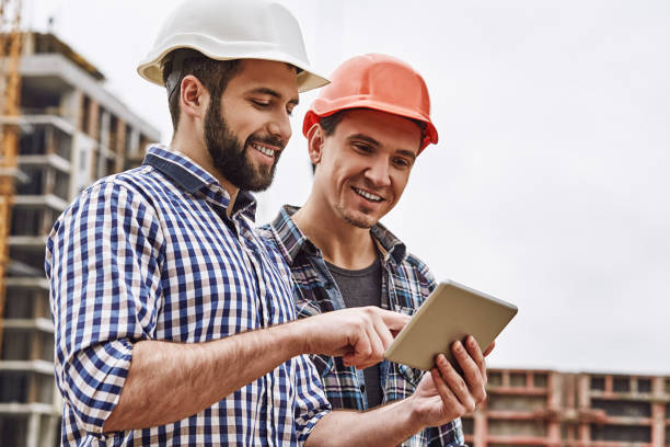 working in team. two young and cheerful builders in protective helmets are using digital tablet and working while standing at construction site - building contractor engineer digital tablet construction imagens e fotografias de stock