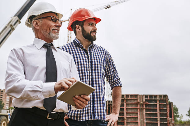 checking work. senior building inspector in formal wear and white helmet is holding a digital tablet and looking with young builder what is already done while standing at construction site. - building contractor engineer digital tablet construction imagens e fotografias de stock