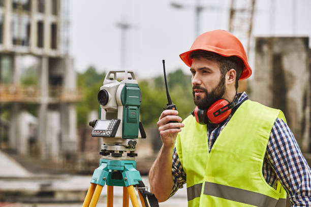 precyzyjne pomiary. inżynier geodeta w odzieży ochronnej i czerwony kask przy użyciu sprzętu geodezyjnego i rozmowy przez walkie talkie stojąc na placu budowy. profesjonalny sprzęt. - azimuth zdjęcia i obrazy z banku zdjęć