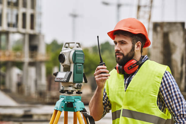 precise measurements. surveyor engineer in protective wear and red helmet using geodetic equipment and talking by walkie talkie while standing at construction site. professional equipment. - azimuth imagens e fotografias de stock