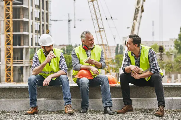 Time for a break. Group of builders in working uniform are eating sandwiches and talking while sitting on stone surface against construction site. Building concept. Lunch concept. Building concept