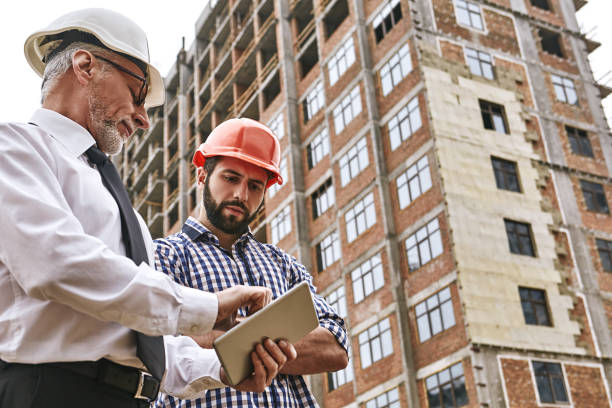 discussion of the work plan. senior professional engineer in formal wear and white helmet is showing project plan on digital tablet to young builder while standing at construction site. - building contractor engineer digital tablet construction imagens e fotografias de stock