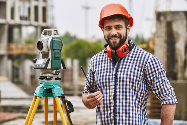 istock Surveyor equipment. Surveyor engineer in protective wear and red helmet using geodetic equipment, holding walkie talkie and smiling while standing at construction site 1142818956