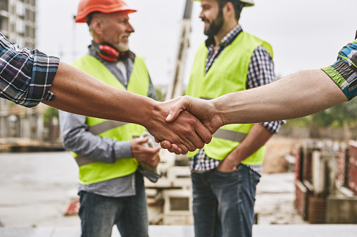 We did it! Close up photo of builders shaking hands against cheerful colleagues while working together at construction site. Team work. Construction concept. Building concept