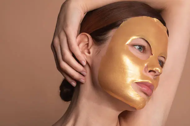 Young woman standing with golden sheet mask on her face and thoughtfully looking away while having one hand on her head