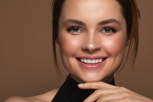 Portrait of a pretty smiling lady with makeup standing isolated on the brown background with her hand under the chin and smiling