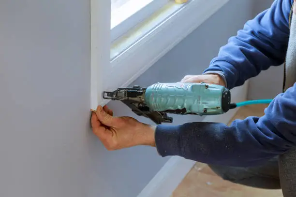 Carpenter using air nail gun to moldings for window, close up on hand and nail gun