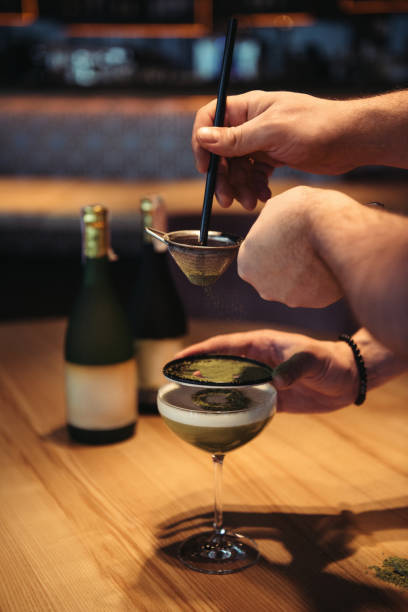 partial view of bartenders preparing alcoholic cocktail using sieve on wooden table in bar