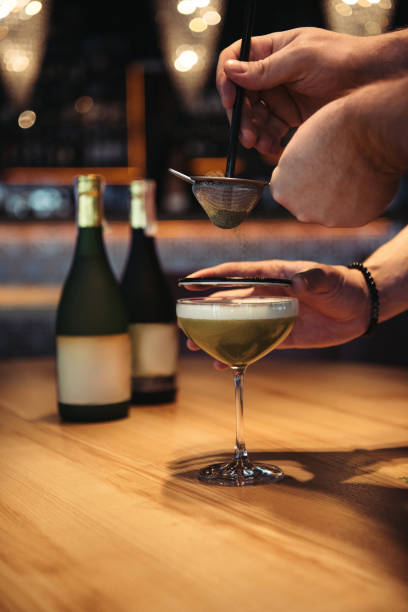 partial view of bartenders preparing alcoholic cocktail using sieve on wooden table in bar