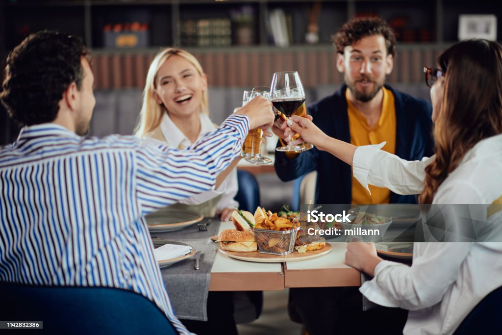 Friends making a toast in restaurant. Four happy muliethnic freinds dressed smart casual cheering with alcohol while sitting at restaurant. Beer - Alcohol Stock Photo