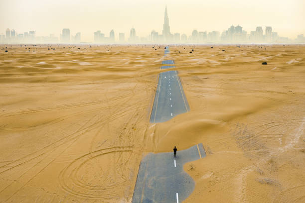 blick von oben, atemberaubende luftaufnahme einer nicht identifizierten person, die auf einer verlassenen straße spaziert, die von sanddünen mitten in der wüste von dubai bedeckt ist.  dubai, vereinigte arabische emirate. - fog desert arabia sunset stock-fotos und bilder