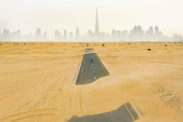 atemberaubende luftaufnahme einer nicht identifizierten person, die auf einer verlassenen straße spaziert, die von sanddünen bedeckt ist, in der wüste von dubai. dubai skyline umgeben von nebel im hintergrund. dubai, vereinigte arabische emirate. - fog desert arabia sunset stock-fotos und bilder