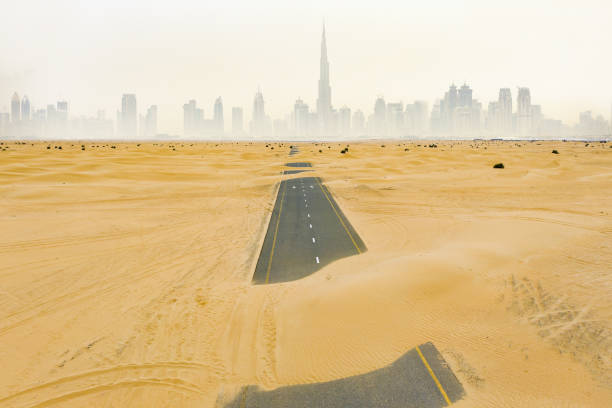 view from above, stunning aerial view of a deserted road covered by sand dunes in the middle of the dubai desert. beautiful dubai skyline surrounded by fog in the background. dubai, united arab emirates. - fog desert arabia sunset imagens e fotografias de stock