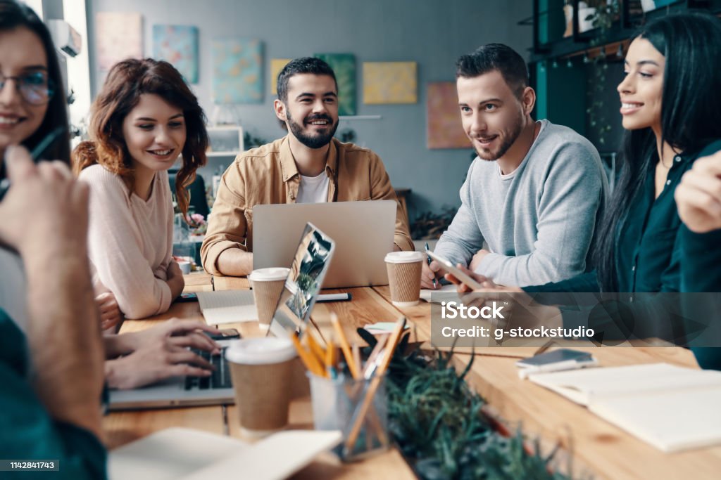 Sharing fresh ideas. Group of young modern people in smart casual wear discussing something and smiling while working in the creative office Staff Meeting Stock Photo